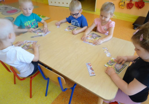 Pięcioro dzieci siedzi przy stole i składa z części obrazek łąki.